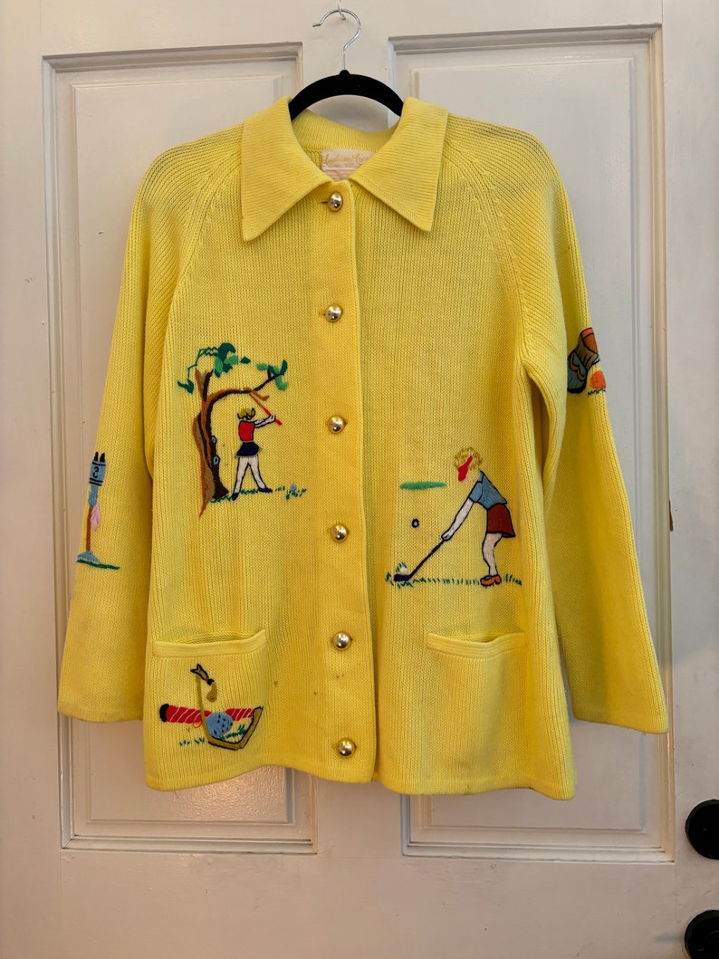May include: A bright yellow cardigan with a collared neckline and gold buttons. The sweater features embroidered golf-themed designs, including golfers, trees, and golf clubs. It has two front pockets and long sleeves.