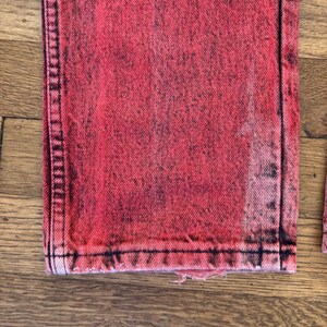 May include: Close-up of a pair of red acid wash jeans. The jeans have a distressed look with black accents and a frayed hem. The fabric has a textured appearance.