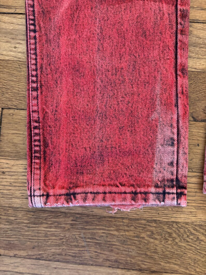 May include: Close-up of a pair of red acid wash jeans. The jeans have a distressed look with black accents and a frayed hem. The fabric has a textured appearance.