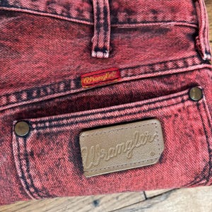 May include: Close-up of red acid wash denim jeans. The jeans feature a brown leather patch with the Wrangler logo on the back pocket and a red Wrangler tag. The jeans have a classic five-pocket design.