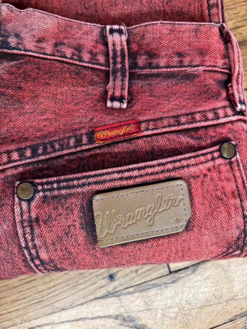 May include: Close-up of red acid wash denim jeans. The jeans feature a brown leather patch with the Wrangler logo on the back pocket and a red Wrangler tag. The jeans have a classic five-pocket design.