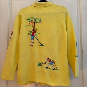 May include: A yellow cardigan sweater with embroidered golf-themed designs. The back features a golfer, tree, and green landscape. The sleeves have additional golf-themed embroidery. The sweater has a collared neckline and button closure.