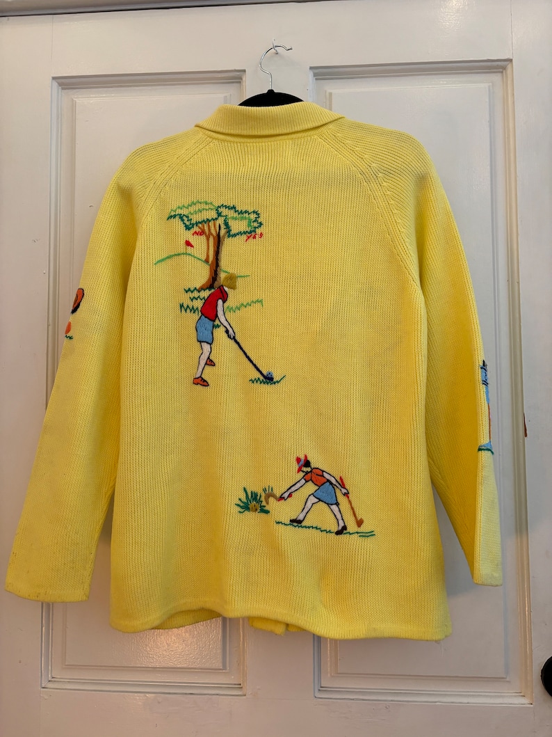 May include: A yellow cardigan sweater with embroidered golf-themed designs. The back features a golfer, tree, and green landscape. The sleeves have additional golf-themed embroidery. The sweater has a collared neckline and button closure.