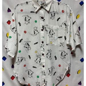 K&ouml;nnte beinhalten: Wei&szlig;es Button-Down-Hemd mit verspieltem Katzenmuster. Das kurz&auml;rmelige Hemd hat schwarze, mit Linien gezeichnete Katzen, bunte Kreise und Zickzackmuster. Es hat eine Brusttasche und dunkelbraune Kn&ouml;pfe. Das Hemd ist ein l&auml;ssiger Stil.