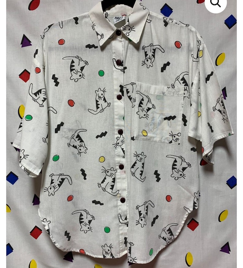 K&ouml;nnte beinhalten: Wei&szlig;es Button-Down-Hemd mit verspieltem Katzenmuster. Das kurz&auml;rmelige Hemd hat schwarze, mit Linien gezeichnete Katzen, bunte Kreise und Zickzackmuster. Es hat eine Brusttasche und dunkelbraune Kn&ouml;pfe. Das Hemd ist ein l&auml;ssiger Stil.