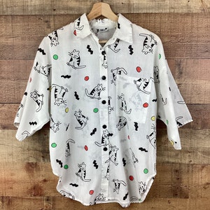 K&ouml;nnte beinhalten: Wei&szlig;es Button-Down-Hemd mit einem verspielten Katzenmuster. Das Hemd hat schwarze Katzen mit gestreiften Schw&auml;nzen, schwarze Zickzackmuster und bunte Punkte. Es hat einen Kragen, eine Brusttasche und schwarze Kn&ouml;pfe.