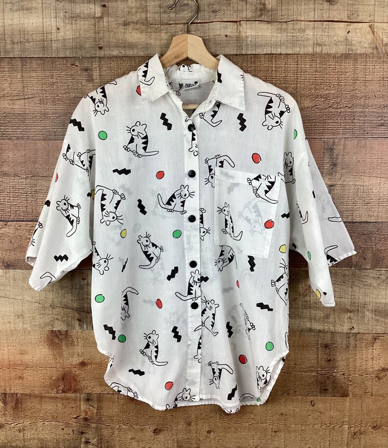 K&ouml;nnte beinhalten: Wei&szlig;es Button-Down-Hemd mit einem verspielten Katzenmuster. Das Hemd hat schwarze Katzen mit gestreiften Schw&auml;nzen, schwarze Zickzackmuster und bunte Punkte. Es hat einen Kragen, eine Brusttasche und schwarze Kn&ouml;pfe.