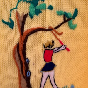 May include: Yellow knit sweater with an embroidered scene of a figure swinging a red club near a tree. The tree has brown and tan bark with green and teal leaves. The figure is wearing a red top, blue skirt, and white pants.