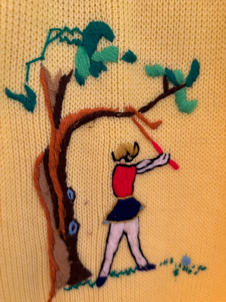 May include: Yellow knit sweater with an embroidered scene of a figure swinging a red club near a tree. The tree has brown and tan bark with green and teal leaves. The figure is wearing a red top, blue skirt, and white pants.