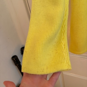 May include: Close-up of a bright yellow knit sleeve. The sleeve has a ribbed texture and a slight flare at the end. The fabric shows some minor imperfections. The background is a white door.