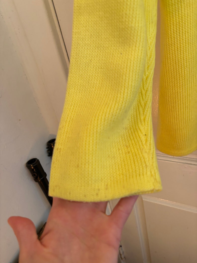 May include: Close-up of a bright yellow knit sleeve. The sleeve has a ribbed texture and a slight flare at the end. The fabric shows some minor imperfections. The background is a white door.