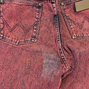 May include: Close-up of the back of red Wrangler jeans. The jeans have two back pockets with the Wrangler logo on a brown leather patch. The stitching is black, and the jeans have a faded, distressed look.