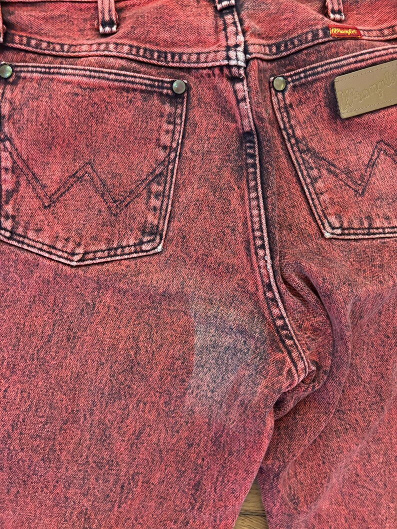 May include: Close-up of the back of red Wrangler jeans. The jeans have two back pockets with the Wrangler logo on a brown leather patch. The stitching is black, and the jeans have a faded, distressed look.