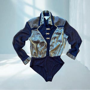 以下が含まれることがあります： ネイビーブルーのボディスーツ。長袖で、メタリックシルバーとゴールドのフロントパネルが付いています。襟付きで、ゴールドカラーのボタンが付いています。袖口は濃い青色です。