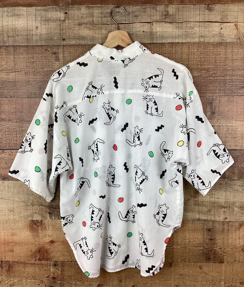 K&ouml;nnte beinhalten: Wei&szlig;es Button-up-Hemd mit einem verspielten Katzenmuster. Das Hemd hat ein sich wiederholendes Design aus schwarz-wei&szlig;en Cartoon-Katzen, zusammen mit bunten Punkten und schwarzen Zickzacklinien. Das Hemd h&auml;ngt an einem Holzb&uuml;gel.