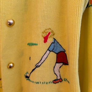 May include: Yellow knit cardigan with gold buttons and embroidered golf-themed design. The embroidery features a golfer in a red skirt and blue shirt, swinging a golf club on a green field. The cardigan has a pocket.