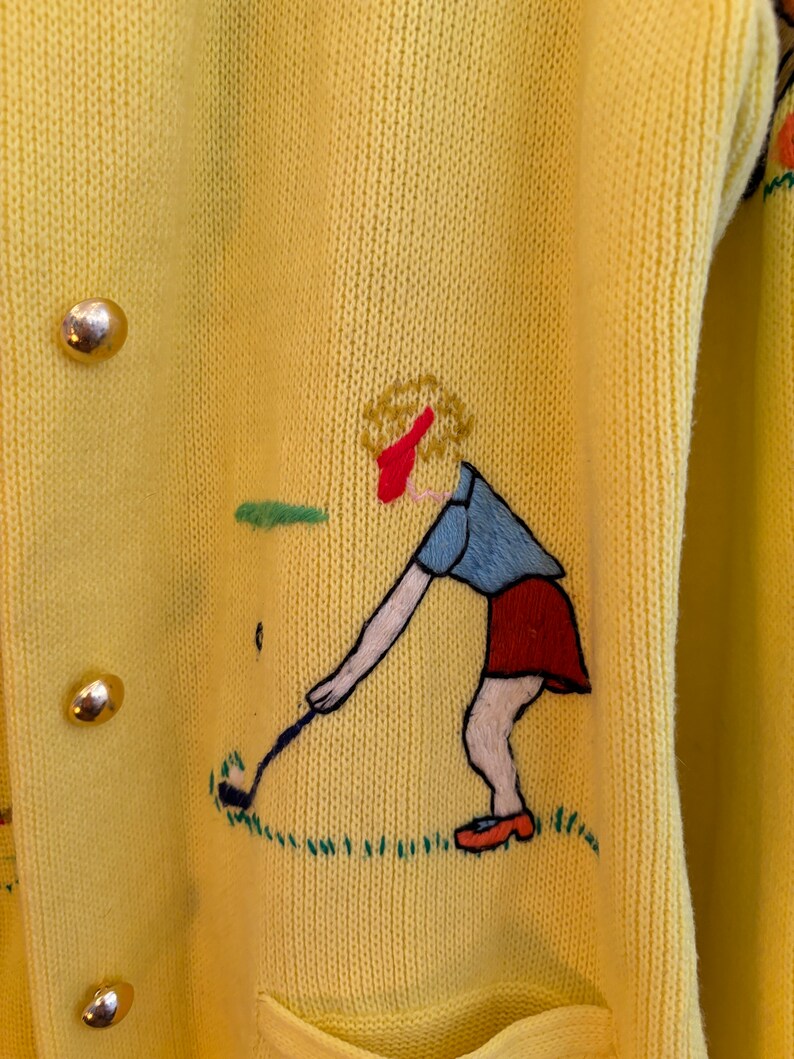 May include: Yellow knit cardigan with gold buttons and embroidered golf-themed design. The embroidery features a golfer in a red skirt and blue shirt, swinging a golf club on a green field. The cardigan has a pocket.