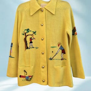May include: A bright yellow cardigan sweater with gold buttons and two front pockets. The sweater features embroidered golf-themed designs, including golfers, trees, and golf clubs. The collar is a classic style, and the sweater is hanging on a black hanger.