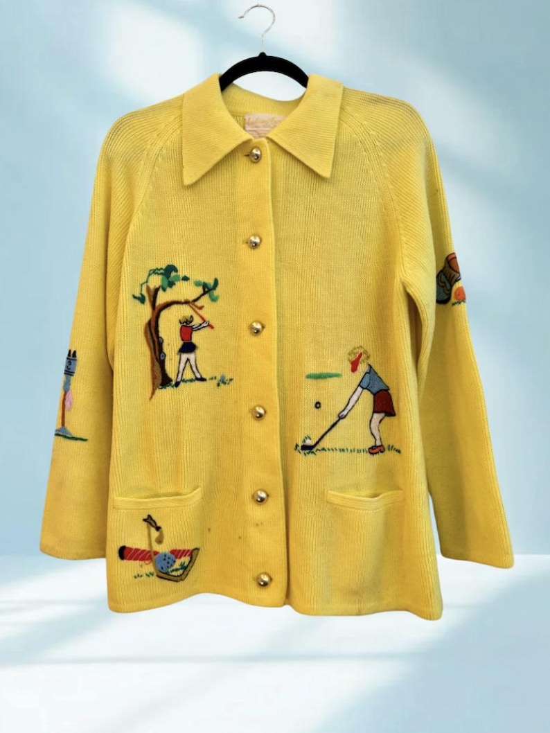 May include: A bright yellow cardigan sweater with gold buttons and two front pockets. The sweater features embroidered golf-themed designs, including golfers, trees, and golf clubs. The collar is a classic style, and the sweater is hanging on a black hanger.