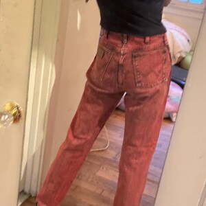 May include: Red denim jeans with a classic five-pocket design. The jeans have a faded, worn look and a straight leg fit. The person in the image is wearing a black t-shirt.