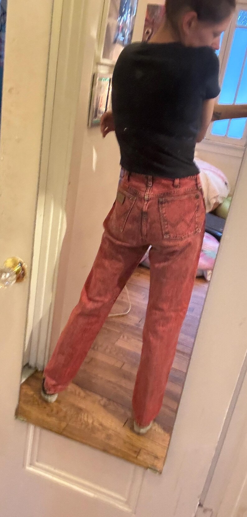 May include: Red denim jeans with a classic five-pocket design. The jeans have a faded, worn look and a straight leg fit. The person in the image is wearing a black t-shirt.