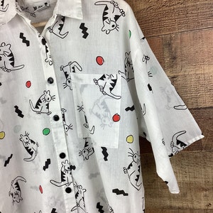 K&ouml;nnte beinhalten: Wei&szlig;es Button-Down-Hemd mit einem verspielten Katzenmuster. Das Hemd zeigt schwarze Katzenillustrationen, schwarze Zickzackformen und bunte Punkte. Es hat eine Brusttasche und schwarze Kn&ouml;pfe. Das Hemd h&auml;ngt an einem Holzkleiderb&uuml;gel.