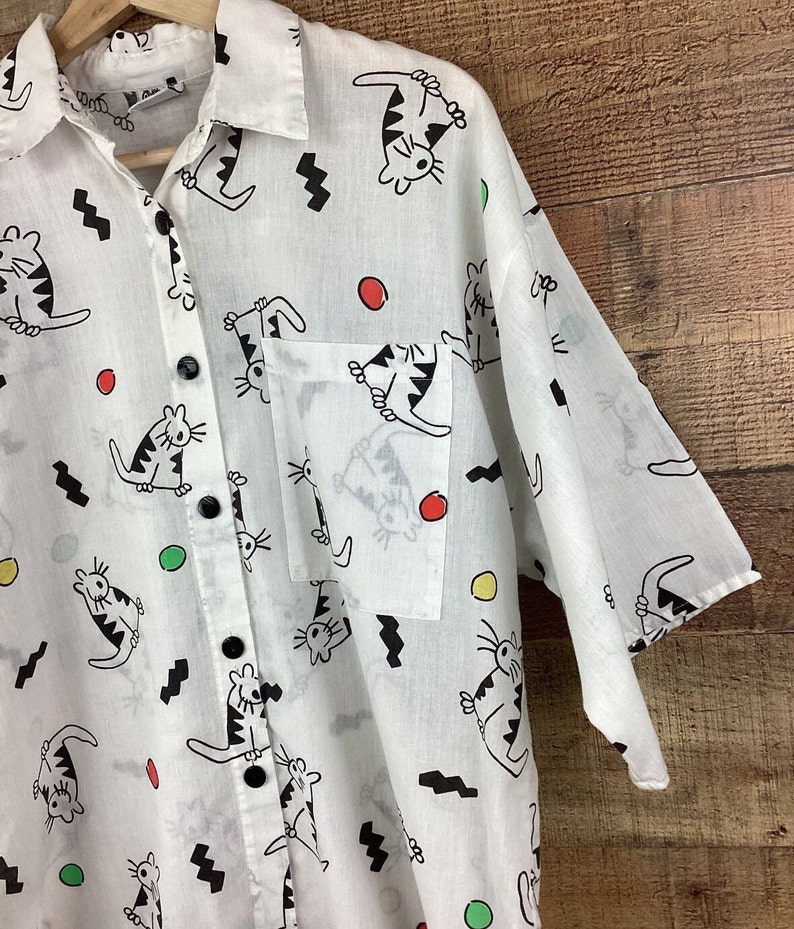K&ouml;nnte beinhalten: Wei&szlig;es Button-Down-Hemd mit einem verspielten Katzenmuster. Das Hemd zeigt schwarze Katzenillustrationen, schwarze Zickzackformen und bunte Punkte. Es hat eine Brusttasche und schwarze Kn&ouml;pfe. Das Hemd h&auml;ngt an einem Holzkleiderb&uuml;gel.