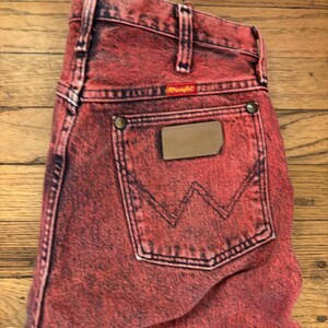 May include: Red acid wash Wrangler jeans. The jeans have a classic five-pocket design, a leather patch, and the Wrangler logo. The back pocket features the iconic 'W' stitching. The jeans are made of a sturdy denim material.