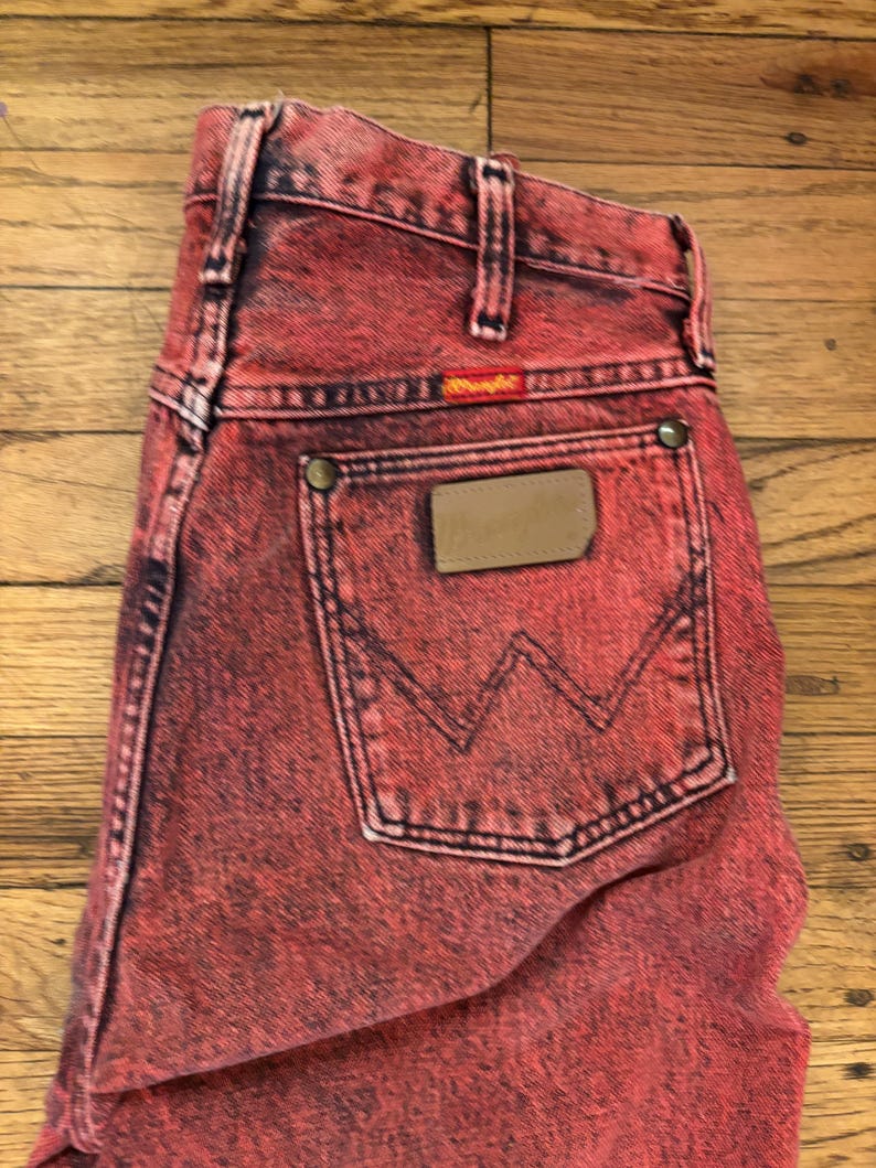 May include: Red acid wash Wrangler jeans. The jeans have a classic five-pocket design, a leather patch, and the Wrangler logo. The back pocket features the iconic 'W' stitching. The jeans are made of a sturdy denim material.
