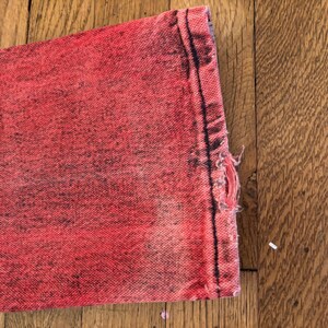 May include: Close-up of a section of red denim jeans with a distressed, faded appearance. The fabric shows signs of wear and tear, with a frayed edge and a small hole. The jeans are on a wooden surface.