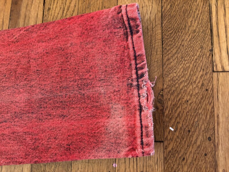 May include: Close-up of a section of red denim jeans with a distressed, faded appearance. The fabric shows signs of wear and tear, with a frayed edge and a small hole. The jeans are on a wooden surface.