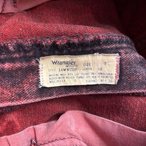 May include: Close-up of red Wrangler jeans. The jeans have a faded, distressed look with black stitching. A tag displays the Wrangler brand, size 9, and length 32. The tag also includes washing instructions and "Made in USA."