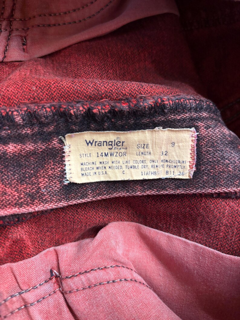 May include: Close-up of red Wrangler jeans. The jeans have a faded, distressed look with black stitching. A tag displays the Wrangler brand, size 9, and length 32. The tag also includes washing instructions and "Made in USA."