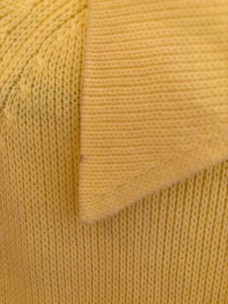 May include: Close-up of a pale yellow knit fabric, possibly a sweater or top. The texture is visible with vertical ribbing and a V-neck collar. The fabric appears soft and comfortable, suitable for casual wear.