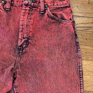 May include: Close-up of red acid wash jeans. The jeans have a high waist, a button and zipper closure, and classic five-pocket styling. The denim is dyed with a mottled effect, creating a unique, vintage look. The jeans are a vibrant red color.