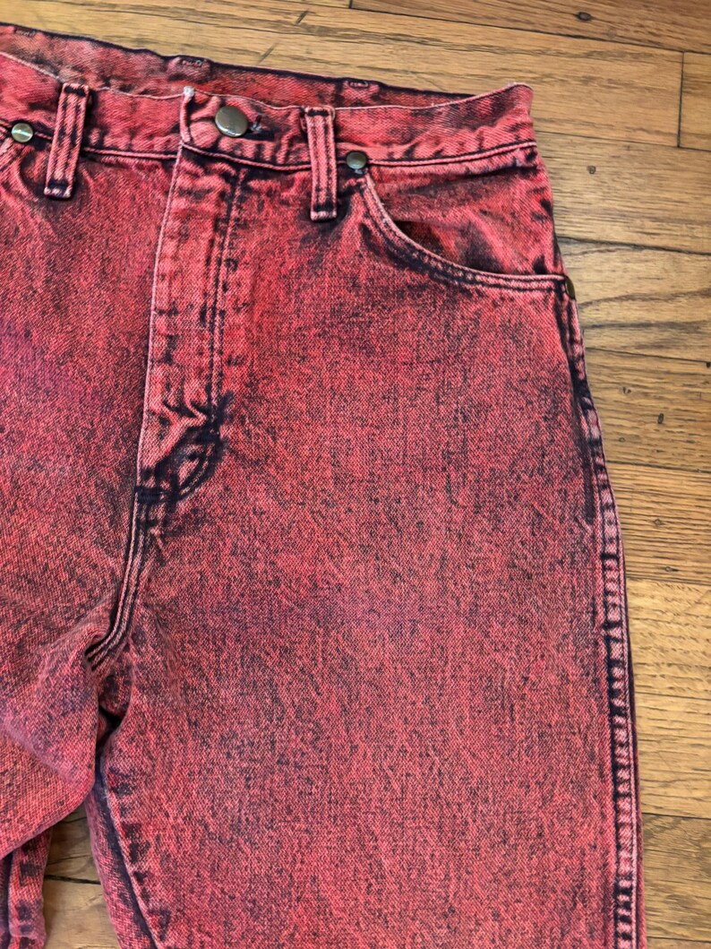 May include: Close-up of red acid wash jeans. The jeans have a high waist, a button and zipper closure, and classic five-pocket styling. The denim is dyed with a mottled effect, creating a unique, vintage look. The jeans are a vibrant red color.