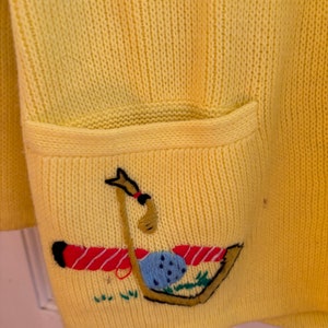 May include: A yellow knit cardigan with a pocket featuring an embroidered golf-themed design. The design includes a golf club, ball, and tee, with red and black accents. The cardigan is made of a soft, textured knit fabric.