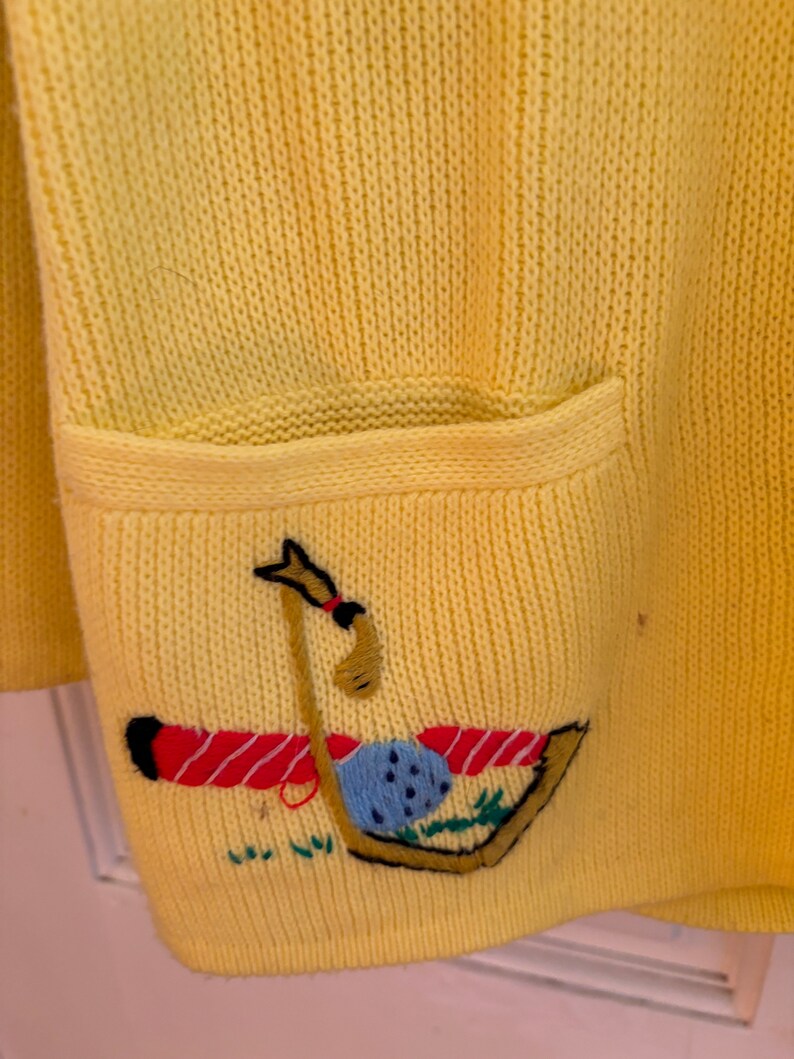 May include: A yellow knit cardigan with a pocket featuring an embroidered golf-themed design. The design includes a golf club, ball, and tee, with red and black accents. The cardigan is made of a soft, textured knit fabric.