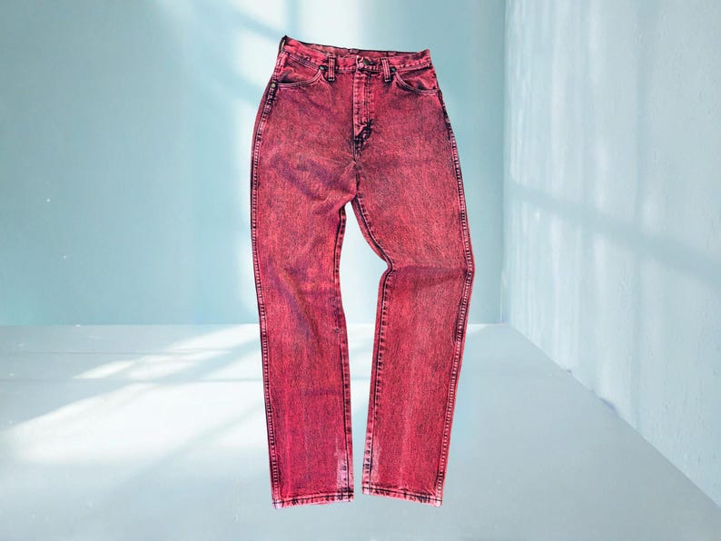 May include: A pair of acid-wash red denim jeans. The jeans have a high waist and a straight leg cut. The denim is a vibrant red with darker red accents, and the jeans are displayed against a light blue background.