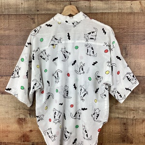 K&ouml;nnte beinhalten: Wei&szlig;es Button-up-Shirt mit einem verspielten Katzenmuster. Das Shirt zeigt schwarze Strichzeichnungen von Katzen in verschiedenen Posen, zusammen mit bunten Tupfen und schwarzen Zickzacklinien. Das Shirt h&auml;ngt an einem Holzkleiderb&uuml;gel.