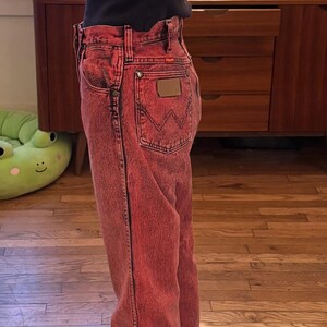 May include: Red acid wash Wrangler jeans with a brown leather patch on the back pocket. The jeans are paired with a black top and checkered slip-on shoes. The jeans have a classic five-pocket design.