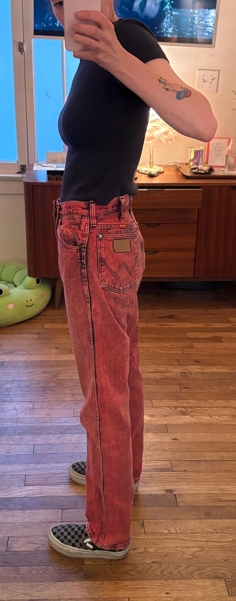 May include: Red acid wash Wrangler jeans with a brown leather patch on the back pocket. The jeans are paired with a black top and checkered slip-on shoes. The jeans have a classic five-pocket design.