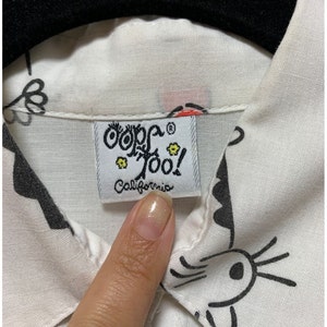 K&ouml;nnte beinhalten: Nahaufnahme eines wei&szlig;en Hemdes mit Kragen und einem schwarz-wei&szlig;en Cartoon-Design. Das Hemd hat ein Etikett mit der Aufschrift "Oops Too! California". Das Etikett ist rechteckig mit wei&szlig;em Hintergrund und schwarzem Text.