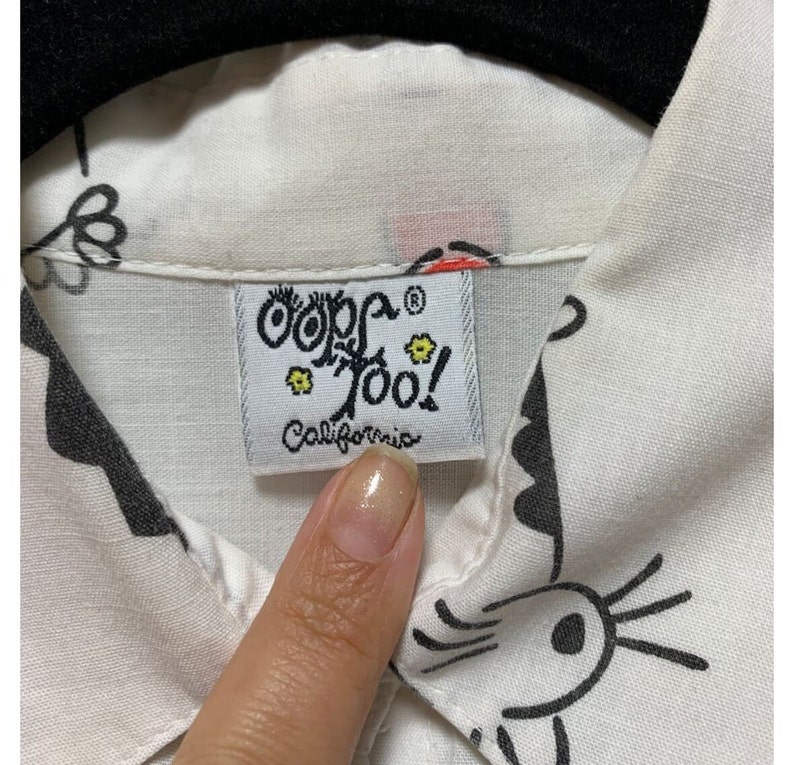 K&ouml;nnte beinhalten: Nahaufnahme eines wei&szlig;en Hemdes mit Kragen und einem schwarz-wei&szlig;en Cartoon-Design. Das Hemd hat ein Etikett mit der Aufschrift "Oops Too! California". Das Etikett ist rechteckig mit wei&szlig;em Hintergrund und schwarzem Text.