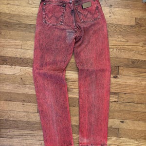May include: Acid wash red denim jeans with the Wrangler logo on the back pockets. The jeans have a faded, distressed look, with a brown leather patch. The jeans are laid flat on a wooden surface.