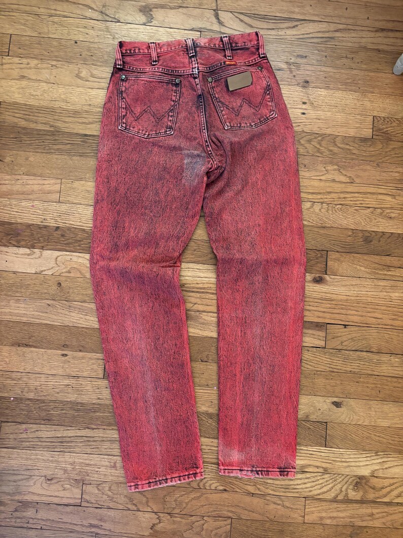 May include: Acid wash red denim jeans with the Wrangler logo on the back pockets. The jeans have a faded, distressed look, with a brown leather patch. The jeans are laid flat on a wooden surface.