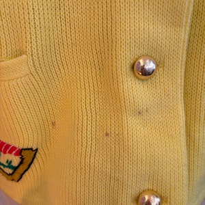 May include: Close-up of a yellow knit cardigan with gold-colored buttons. The cardigan has a pocket and an embroidered design on the left side. The knit fabric has a textured appearance.