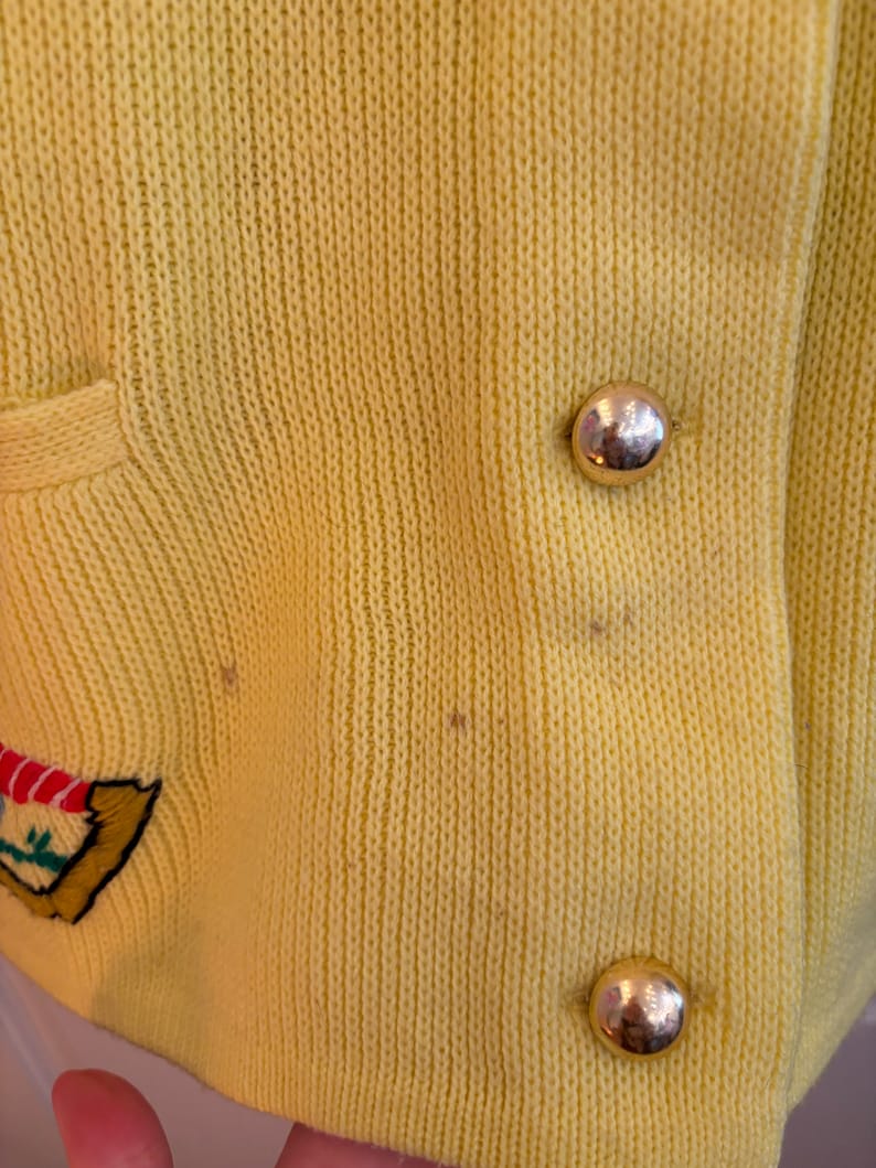May include: Close-up of a yellow knit cardigan with gold-colored buttons. The cardigan has a pocket and an embroidered design on the left side. The knit fabric has a textured appearance.