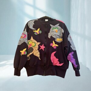 May include: Black zip-up jacket with colorful owl and star appliqués. The jacket features a whimsical design with various owl figures, stars, and moon shapes in shades of gray, pink, yellow, and purple. The jacket is hanging on a hanger.