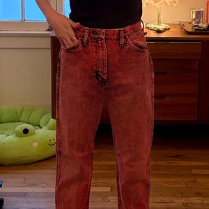 May include: A person wearing a black t-shirt and red acid wash jeans. The person is also wearing black and gray checkered slip-on shoes. The background includes a window and a wooden floor.